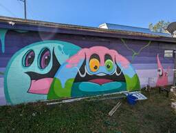 Patch Whisky is a well-known street artist. This is his studio in North Charleston, South Carolina, located at 4431 Spruill Avenue. The mural depicts two pink creatures in the bottom right, two large and colorful smiling monsters in the bottom center, and a patterned purple background. 