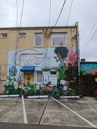 Alycia Alley runs behind a row of businesses in the artsy neighborhood of Avondale, West Ashley, Charleston. This artwork was photographed from Alycia Alley and painted by an unknown artist on the rear wall of Charleston Yoga Massage. It depicts a black woman with an afro perched halfway up a large flower stem blowing a handful of seeds over a nature scene below her. The nature scene contains flowers and mushrooms, and a brown bag labeled "SEED." 