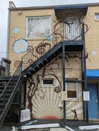 Alycia Alley runs behind a row of businesses in the artsy neighborhood Avondale, West Ashley, Charleston. This artwork was photographed from Alycia Alley and painted by an unknown artist on the rear wall of Charleston Yoga Massage. It depicts a rising/setting sun with a silhouetted man carrying a fishing pole across the bottom, and a brown tree following the path of the staircase toward the top of the building. 