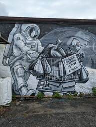 This mural depicts a black and grey scene of an individual in a space suit pushing a shopping cart on the surface of a non-Earth celestial body. The cart is full of bags and various items and has a sign on the front reading "global warming is just a hoax." The s of "is" was replaced by a dollar sign, and the t of "just" was replaced by a Christian cross. 