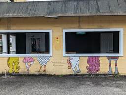 This mural by @tami_draws is located on a side wall of Gene's Haufbrau facing the parking lot. It is an interactive art piece with the lower halves of six creatures (dinosaur, ballerina, chicken, unicorn, octopus, and robot) set underneath two window openings. The effect is that if one were to stand on the other side of the window, it would look like your bottom half was replaced by one of the creatures. 