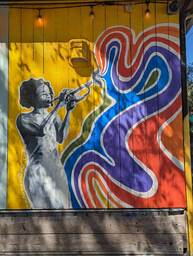 The Charleston Pour House is a live music venue located at 1977 Maybank Highway in James Island, SC. This mural by @mia_loia has a black and grey woman playing a trumpet, and out of the trumpet five brightly colored wavy lines emerge. The background is solid yellow. 