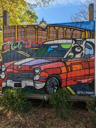 The Charleston Pour House is a live music venue located at 1977 Maybank Highway in James Island, SC. This mural by @m.leilaniart shows a raccoon looking out of the driver's window of a red car with a street and wall in the background. 