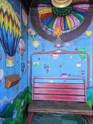 The Charleston Pour House is a live music venue located at 1977 Maybank Highway in James Island, SC. This mural by Leslie Caneda depicts hot air balloons floating on a blue background. The bench in front of the mural is made to look like its being lifted into the air by a hot air balloon.